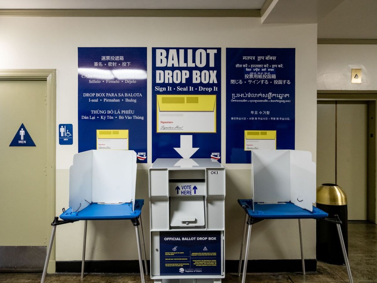 A large metal ballot drop box, similar to a mailbox, with the words "ballot drop box" written on a sign over it, flanked by two voting booths.