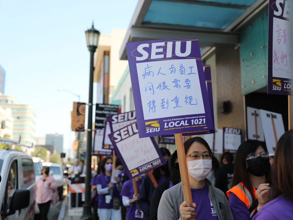 A young Asian woman with a medical mask hold an SEIU picket sign with Chinese characters. Other workers carrying signs can be seen walking on the city sidewalk behind her.