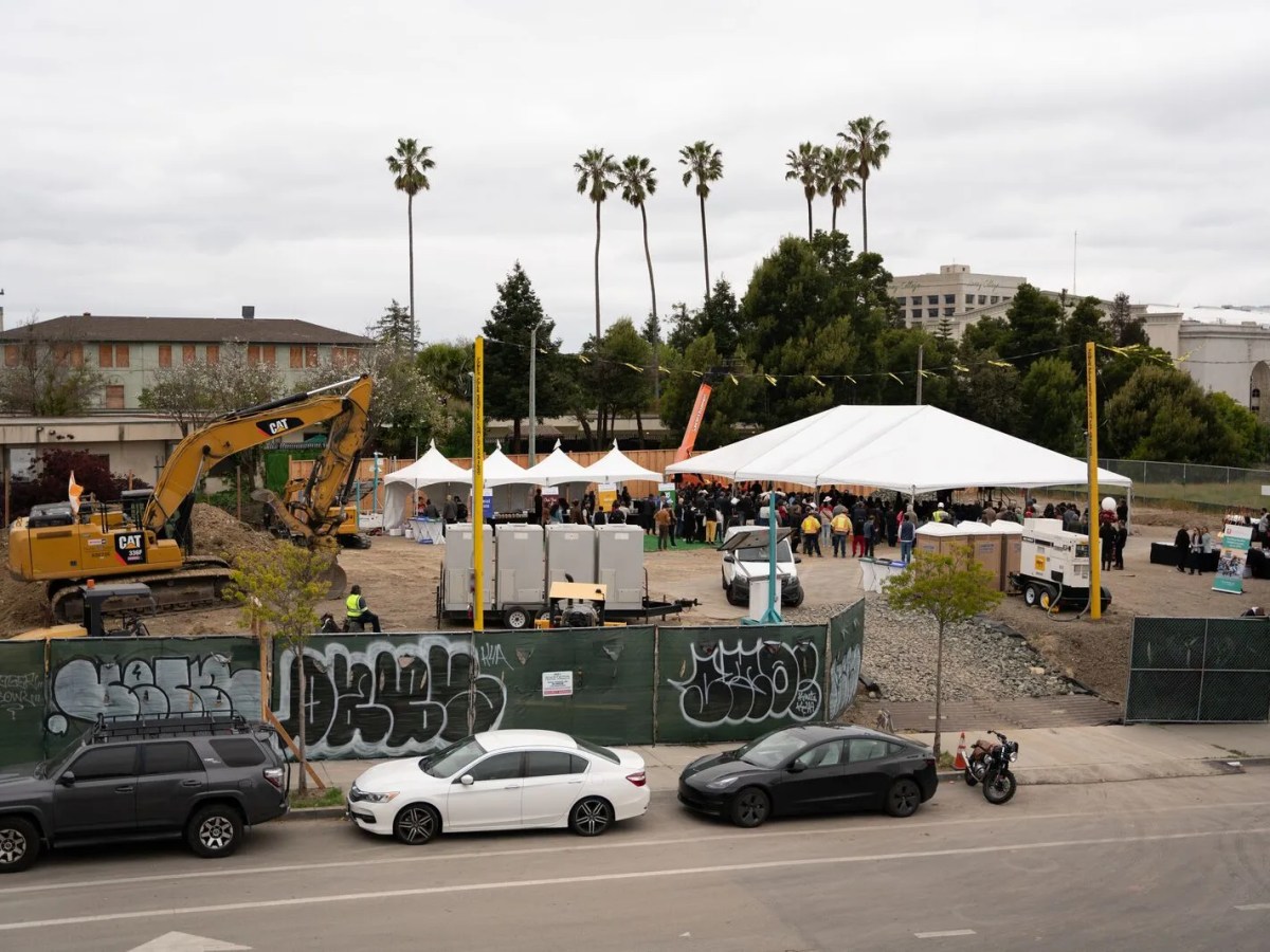 Big construction site off a city street, with people milling about and palm trees and buildings in the background
