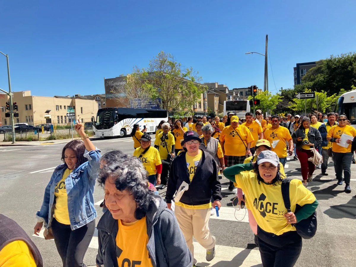 A large group of people in matching yellow shirts crosses a street on a sunny day. One has a fist raised.