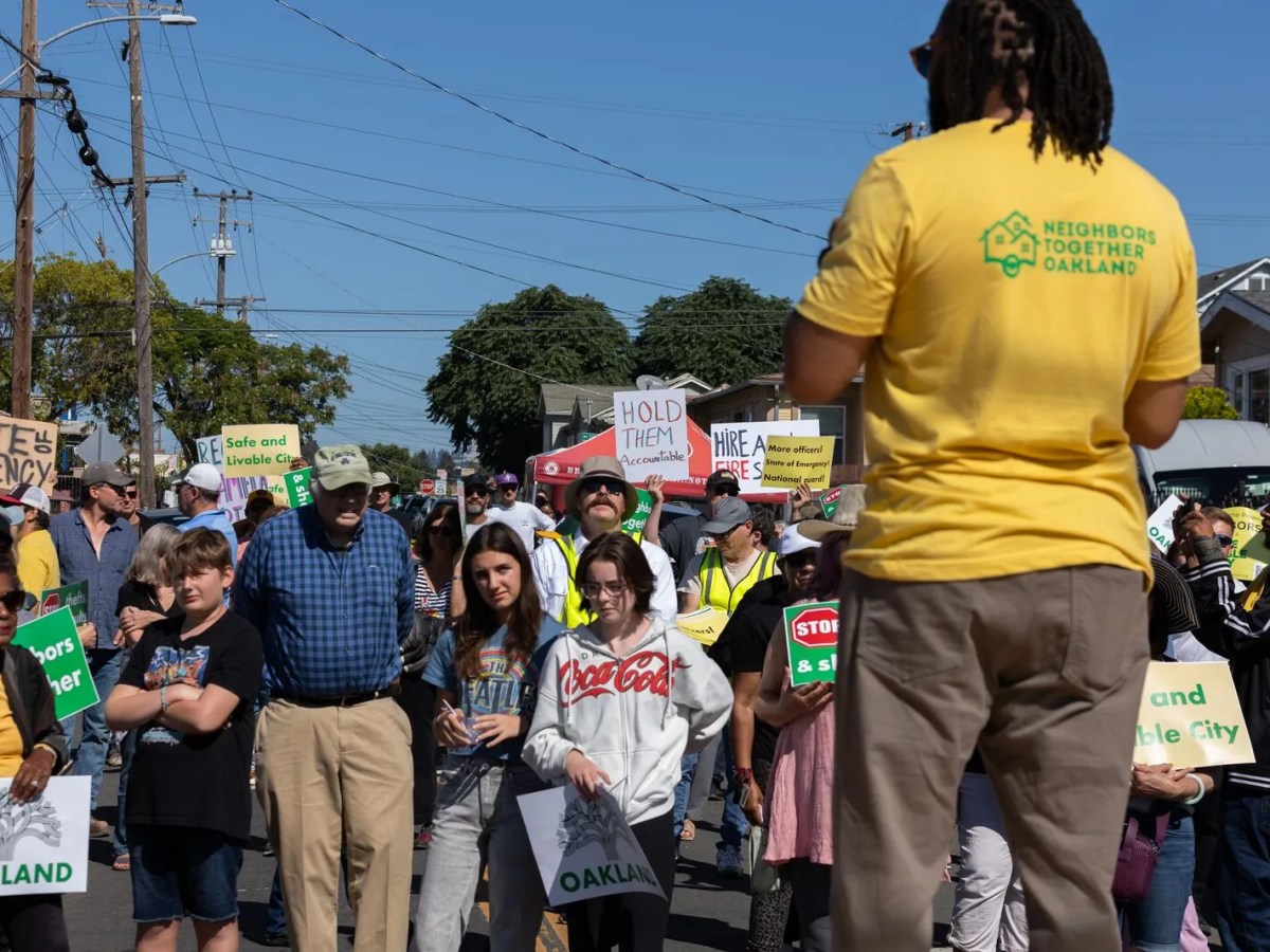 A man in a shirt that says "Neighbors Together Oakland" addresses a diverse crowd of people at a rally.