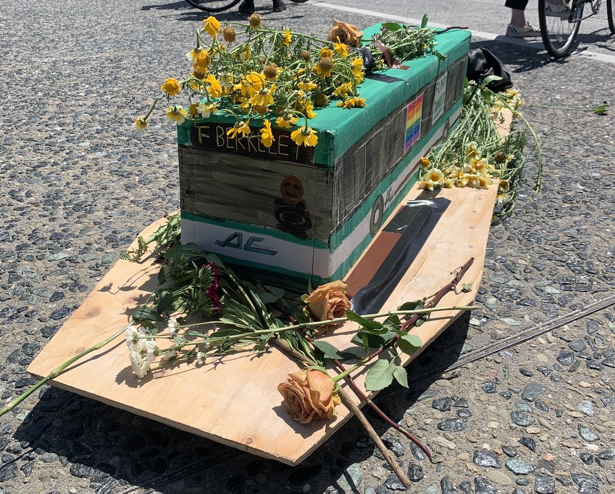 A model of an AC Transit bus made of cardboard, topped and surrounded with flowers, set atop a coffin-shaped board.