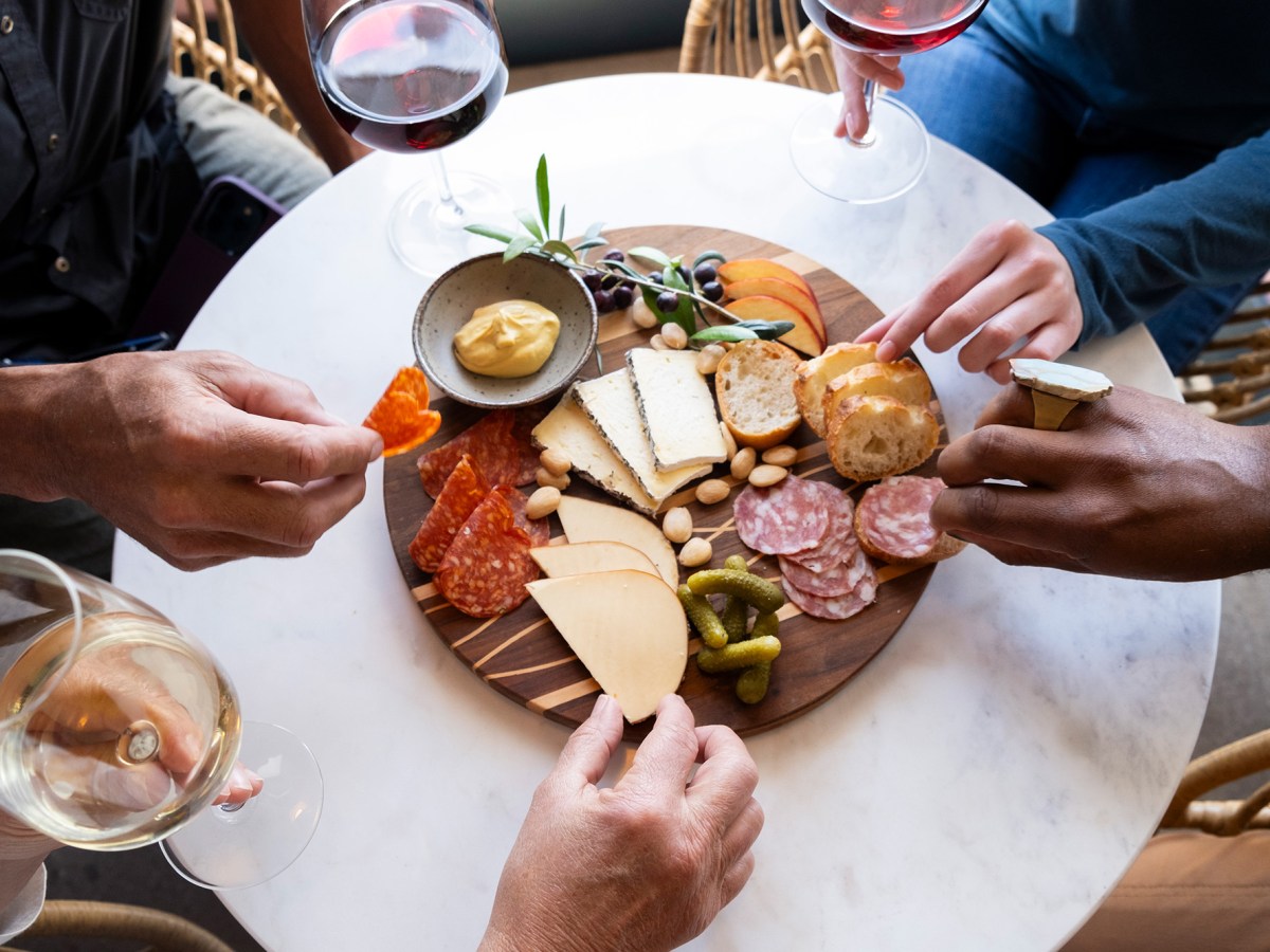 A white round table with a Charcuterie Board on top is surrounded by hands grabbing at different items and wine glasses.