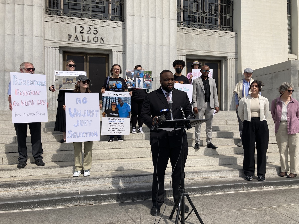 A Black man in a suit stands in front of a diverse group of people holdings signs on the steps of the Rene Davidson Courthouse in Oakland.