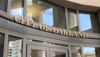 Bronze lettering that spells "City of Oakland" resting above a glass doorway. The address number 150 is above the letters. The sign is surrounded by windows, and above it a sliver of stone wall is visible. Buildings are reflected in the windows.