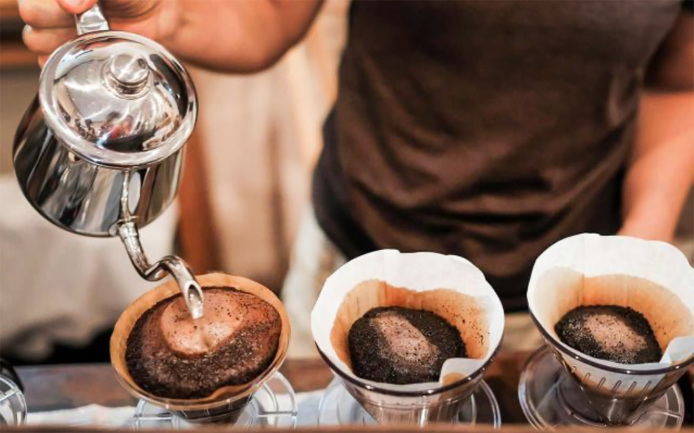 A row of three coffee pour overs and a hand holding a silver pot is adding water to one of them.