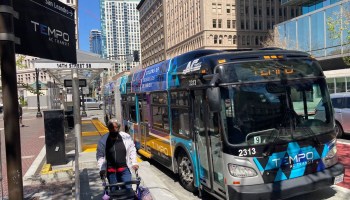 Tall buildings tower over Broadway in the heart of downtown Oakland. A large bus with "TEMPO, AC Transit" written on the front of it drives past a bus stop and a person using a walker.